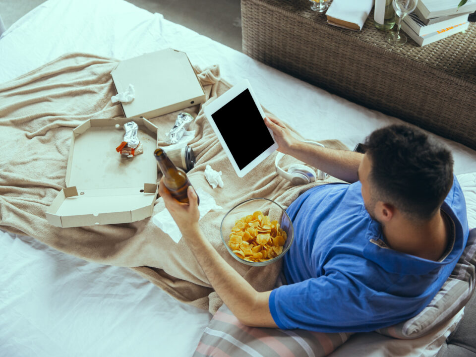 homem sentado na cama com as pernas cobertas com um lençol e caixas de pizza vazias por cima, retratando uma aparência de preguiça e desleixo. Homem está com um tablet em uma mão enquanto na outra, segura uma garrafa de cerveja. Imagem mostra também um pote de suvrilhos perto do homem que usa uma camiseta azul.