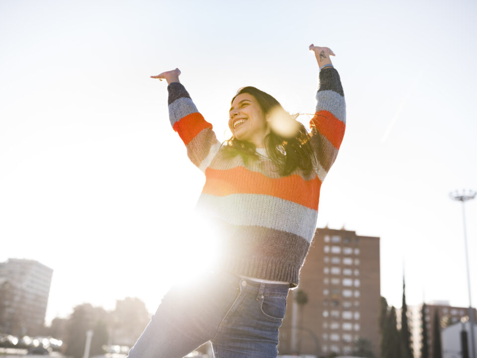 Mulher feliz, com os braços levantados e uma das pernas também, representando energia e alegria. Usa um tricot listrado e uma calça jean, em um ambiente urbano, em frente a prédios.