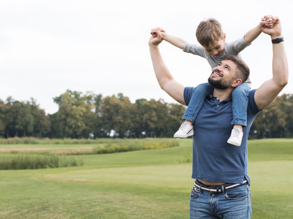 Homem com criança sentada em suas costas feliz sorrindo para a criança. Ele olha pra cima como se tivesse interagindo com a criança. Veste camiseta polo azul e calça jeans e a criança blusa branca, calça jeans e tênis branco.