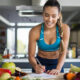 mulher pele morena, de trança longa de lado, com roupa de treino apoiada em uma mesa, escrevendo algo em uma folha. Mesa cheia de frutas e alimentos saudáveis.