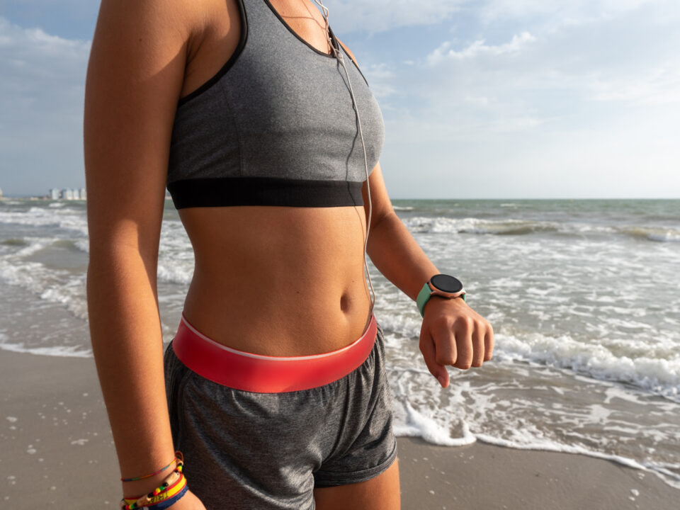 Mulher com top e short de academia, treinando na praia e olhando no relogio.