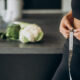 Abdomen de uma mulher na cozinha, medindo a barriga magra com uma fita métrica. Em cima do balcão, algumas verduras