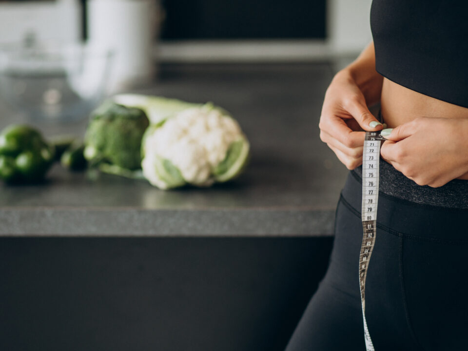 Abdomen de uma mulher na cozinha, medindo a barriga magra com uma fita métrica. Em cima do balcão, algumas verduras