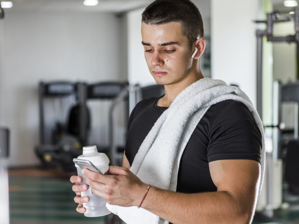 Hoemm com toalha no ombro, segurando uma garrafa de suplementos e olhando o celular, em um ambiente de academia