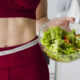 moça com uma roupa de academia segurando um bowl com salada. Ela está com uma roupa vermelha e tem uma fita métrica passando na sua cintura.