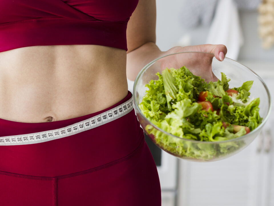 moça com uma roupa de academia segurando um bowl com salada. Ela está com uma roupa vermelha e tem uma fita métrica passando na sua cintura.