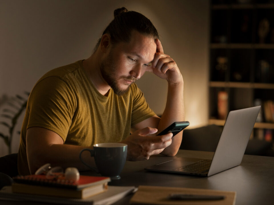 Homem sentado em uma mesa com o celular na mão. Na mesa tem um notebook na frente dele e alguns livros ao seu lado