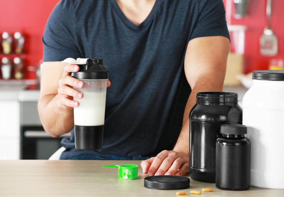 homem segurando uma sheiqueira com potes de suplementos em cima da mesa